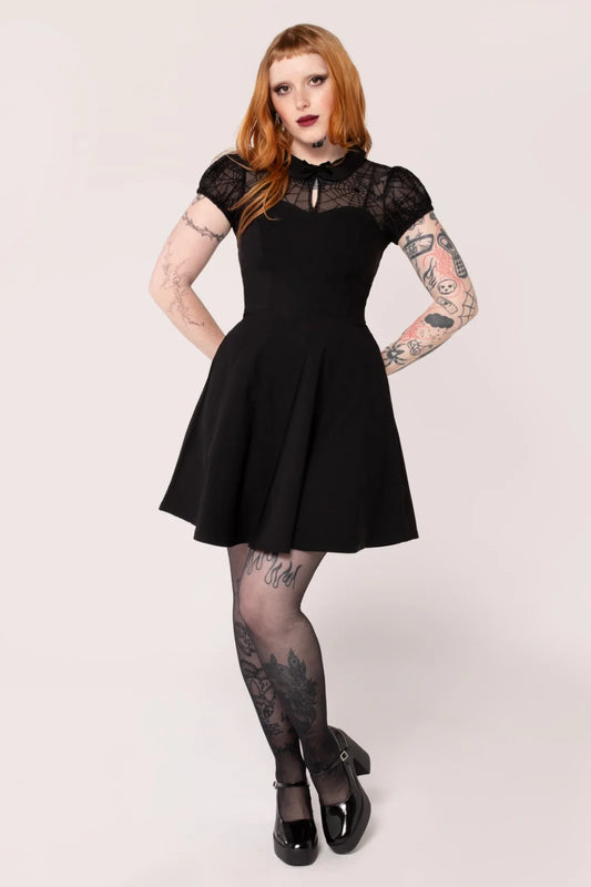 A model wearing a black mini dress with mesh fabric on the chest and sleeves, and velvet flocked spider webs on the chest and waistline. The dress has a sweetheart neckline and a fitted bodice with a skirt that flares out at the waistline.